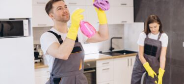 Cleaning company employees wearing gray overalls and yellow gloves cleaning kitchen appliances and floor